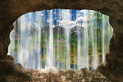 Looking out of a cave under a waterfall onto an idyllic scene in a small river valley. Through a gap in the falling water a cottage is visible just be...